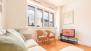 Een moderne woonkamer met een bank, twee rieten stoelen, een televisie en een raam met uitzicht op gebouwen.