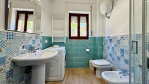 Une salle de bain équipée d’un lavabo, de toilettes et d’un bidet, dont le sol et les murs sont recouverts de carreaux aux motifs bleus et blancs, avec un plancher en bois et une fenêtre à volets.