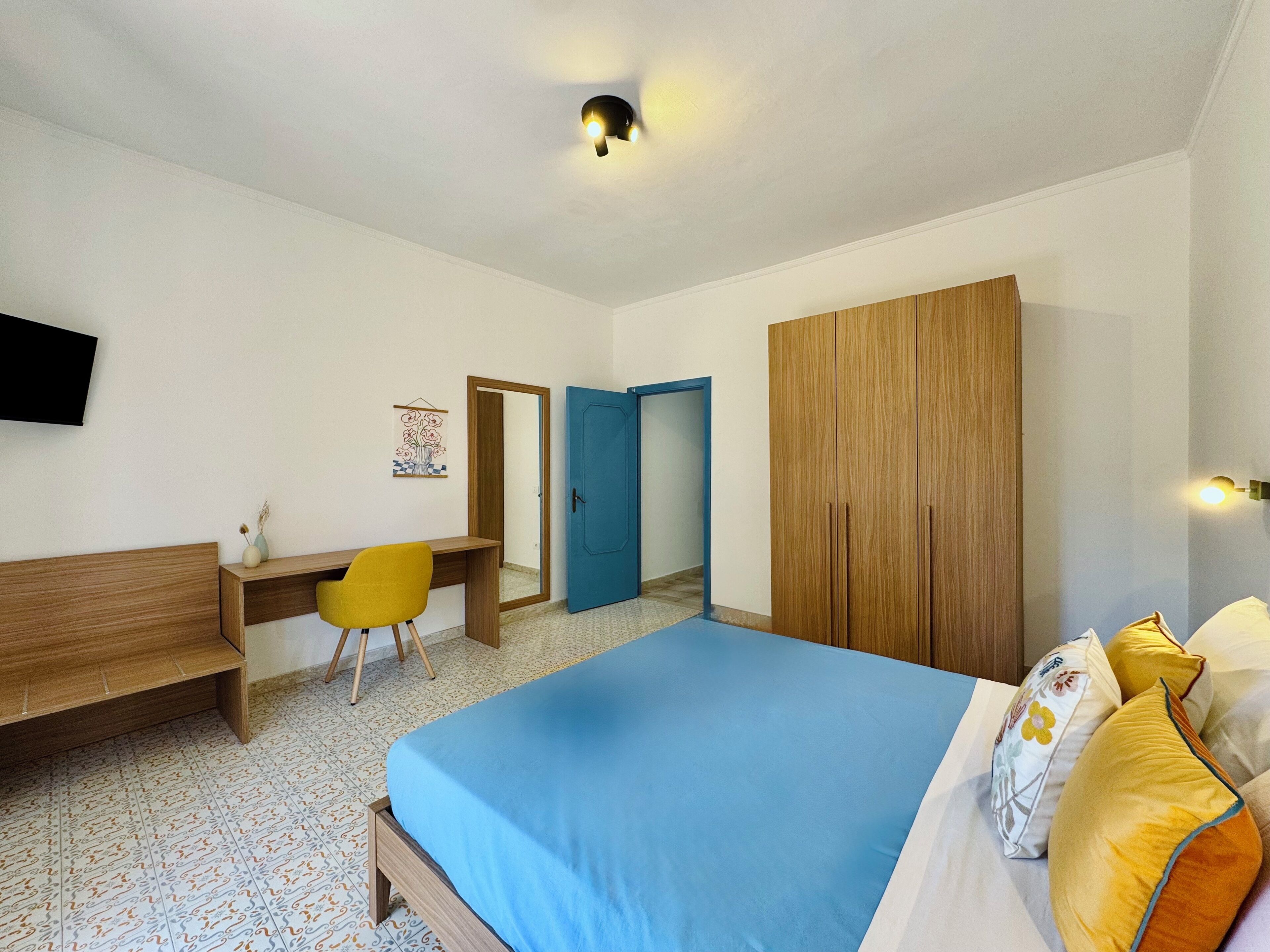 A bedroom with a bed, a desk, a chair, a wardrobe, and a television on the wall.