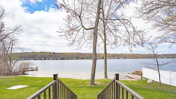 Blick von einer Terrasse auf einen See mit Holzgeländer, einer Rasenfläche und Bäumen im Hintergrund.