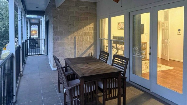 Une terrasse couverte avec une table et des chaises en bois, un ventilateur de plafond et de grandes portes vitrées donnant sur un espace intérieur.
