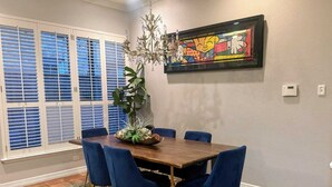 Un coin repas avec une table en bois, des chaises à l’assise rembourrée de couleur bleue, une plante décorative et une œuvre d’art colorée accrochée au mur.