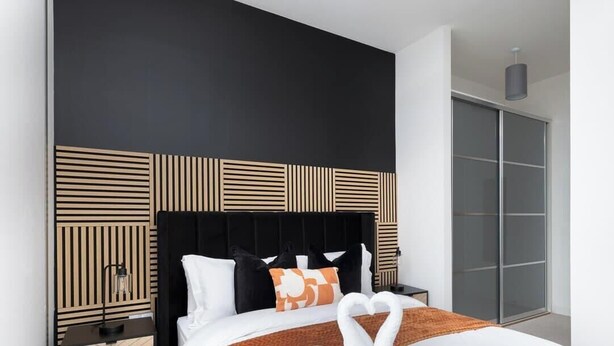 Un dormitorio moderno con cabecera negra, cama con ropa de cama blanca y naranja, y un amplio armario con puerta corredera de vidrio.