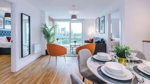 A modern living room with a dining area, large windows, and a city view. - Waterfront 2BR | 2 Bath | Free Gym + Parking (Manchester)