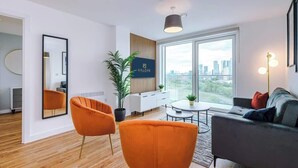A modern living room with a sofa, two orange chairs, a TV, and a large window with a city view. - Waterfront 2BR | 2 Bath | Free Gym + Parking (Manchester)