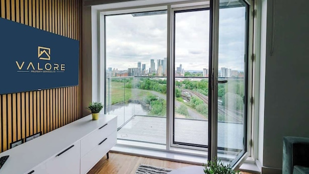 A modern room with a city view, a sliding glass door, and a logo on the wall. - Waterfront 2BR | 2 Bath | Free Gym + Parking (Manchester)