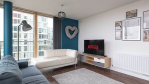 Een moderne woonkamer met een witte bank, een grijze bank, een televisie op een houten dressoir en een hartvormige decoratie aan de muur.