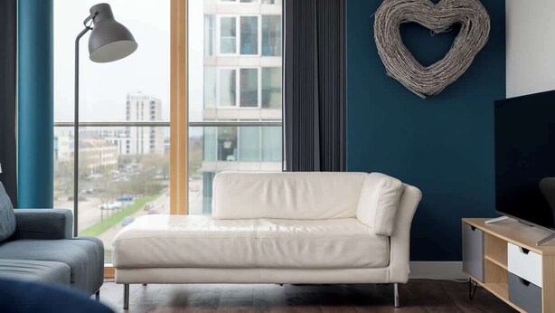 Een moderne woonkamer met een witte bank, een vloerlamp, een hartvormige decoratie aan de muur en uitzicht op gebouwen door grote ramen.