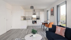 A modern kitchen with white cabinets, a dining area with a round table and chairs, and a living room with a dark sofa and a plant on a coffee table. - Luxury 1Bed| Canal Views|Free Parking|Central MK (Milton Keynes)