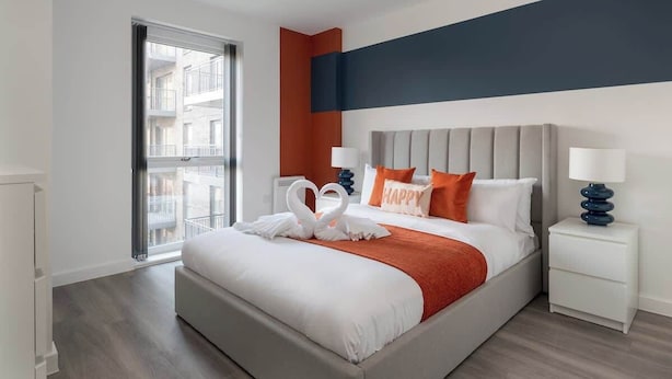A modern hotel room with a large bed, a window with a view of the city, a nightstand with a lamp, and a wall with a blue accent. - Luxury 1Bed| Canal Views|Free Parking|Central MK (Milton Keynes)