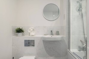 Un baño moderno con cabina de ducha de vidrio, un espejo redondo, lavamanos de mármol y una pequeña planta en maceta.