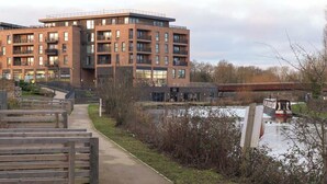 A modern multi-story building with a brick facade, a covered walkway, and a river with a boat. - Luxury 1Bed| Canal Views|Free Parking|Central MK (Milton Keynes)