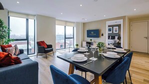 A modern dining area with a large table set for six, a sofa with red cushions, and a city view through large windows.