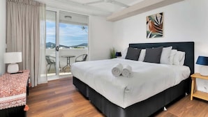 A hotel room with a large bed, a ceiling fan, a view through a sliding glass door, and a small table with a lamp. - Coastal Chic in Heart of Cairns with Rooftop Pool (Cairns)
