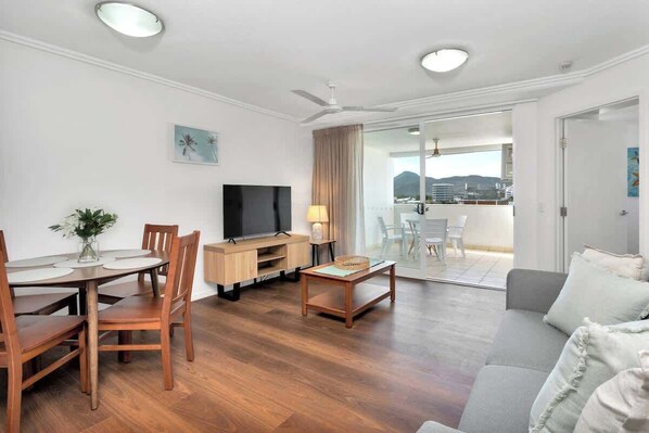 A modern living room with a dining area, a flat-screen TV, a ceiling fan, and a balcony with a view. - Central Plaza: Adjacent Apartments w/ Rooftop Pool (Cairns)