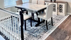 Un espace repas comprenant une table, des chaises, un ventilateur de plafond, une fenêtre avec des stores, un tapis décoratif et un meuble au motif géométrique.