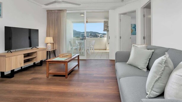 Sala de estar con televisor de pantalla plana, mueble de entretenimiento de madera, una mesa de centro, sofá gris con cojines y una puerta corrediza de vidrio que da a un balcón con vistas.