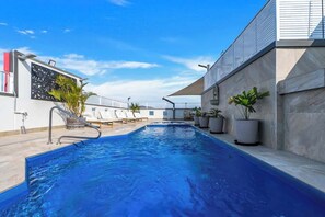 A swimming pool with a metal ladder, lounge chairs, potted plants, and a modern building with a sloped roof.