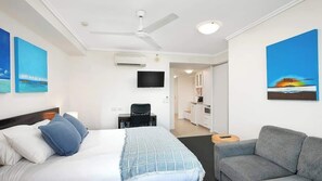 Habitación de hotel con una cama, un sofá gris, un escritorio con una silla, un televisor y obras de arte en las paredes.