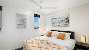 Une chambre d’hôtel avec un lit, un ventilateur de plafond, deux tableaux accrochés au mur, une fenêtre et une table de chevet avec une lampe.