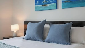 A neatly made bed with two pillows, a bedside lamp, and two paintings of islands on the wall. - Seaside Azul—A Central Studio with Rooftop Pool (Cairns)