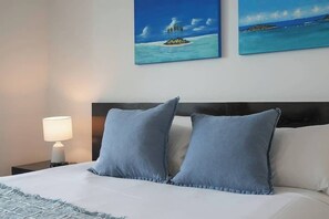 A neatly made bed with two pillows, a bedside lamp, and two paintings of islands on the wall.