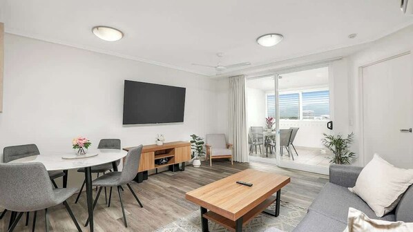A modern living room with a flat-screen TV, grey sofas, a wooden coffee table, and a dining area with a round table and chairs. - Cairns Skyline—Chic Seaside Stay with Rooftop Pool (Cairns)
