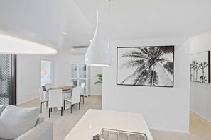 Un salon moderne comprenant un coin repas, un canapé blanc, une table basse et un grand tableau encadré représentant des feuilles de palmier, accroché au mur.