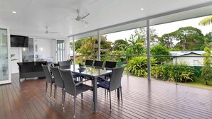 A modern outdoor dining area with a glass table and chairs, a ceiling fan, and a view of lush greenery.
