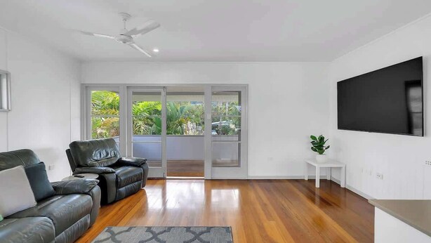A modern living room with a sofa, a coffee table, a ceiling fan, a TV, and a sliding glass door leading to a balcony with greenery.