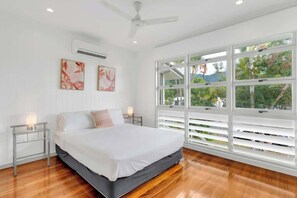Une chambre à coucher avec un lit, des murs blancs, un ventilateur de plafond et de grandes fenêtres équipées de stores.