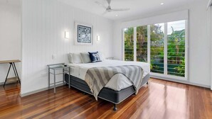 A bedroom with a bed, a desk, a standing lamp, a ceiling fan, and a large window with a view of greenery.