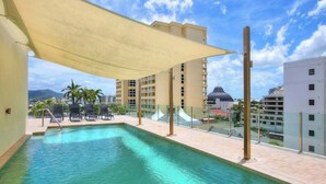 A rooftop swimming pool with a canopy, lounge chairs, and a view of surrounding buildings and palm trees. - Resort-Style Cityview Getaway in Perfect Location (Cairns)