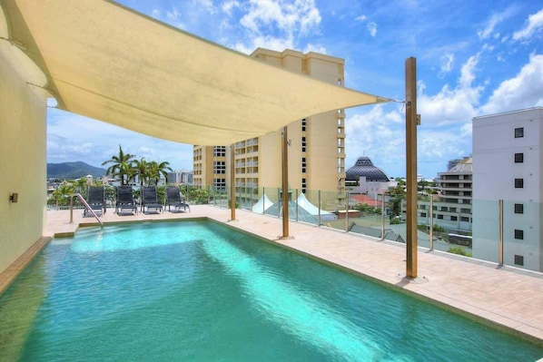 A rooftop swimming pool with a canopy, lounge chairs, and a view of surrounding buildings and palm trees.