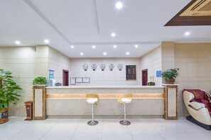 Zona de recepción de hotel con mostrador, dos taburetes de bar, una planta en maceta y un reloj en la pared.