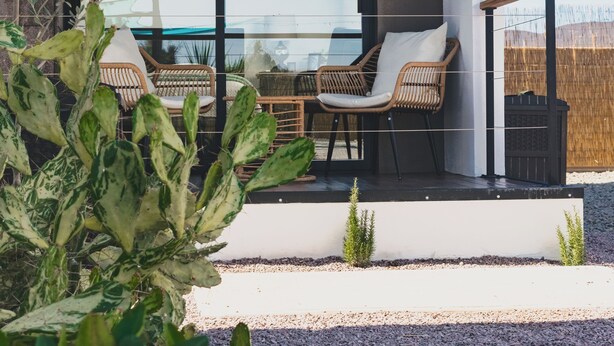 A modern outdoor seating area with wicker chairs and a glass table, surrounded by a gravel path and potted plants.