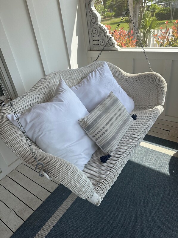 A wicker swing chair with white and striped cushions, hanging by a chain, on a wooden deck with a view of a garden.