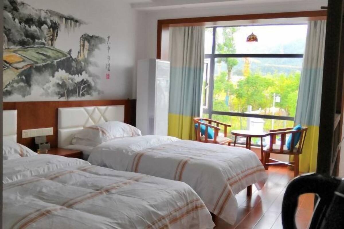 Une chambre d’hôtel avec deux lits, une table à manger, des chaises, une grande fenêtre donnant sur un beau paysage et un tableau accroché au mur.