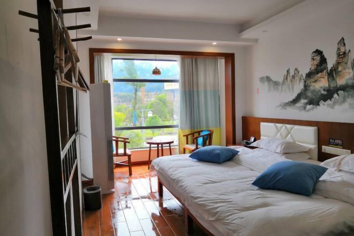 Une chambre d’hôtel avec un lit, un tableau, une fenêtre avec des rideaux et une vue sur la verdure.