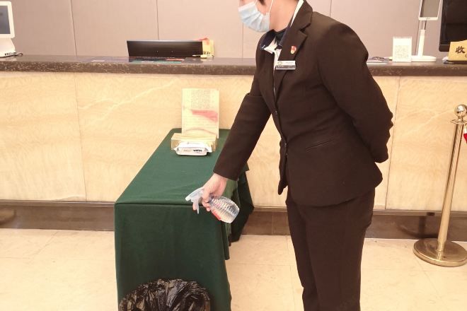 A person in a brown suit standing at a reception desk with a green tablecloth, a computer monitor, and a telephone.