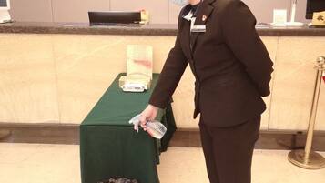 A person in a brown suit standing at a reception desk with a green tablecloth, a computer monitor, and a telephone.