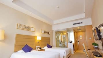 Une chambre d’hôtel avec deux lits, un bureau, une chaise, une télévision et une salle de bain visible à travers une porte en verre.