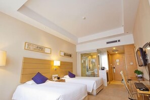 Une chambre d’hôtel avec deux lits, un bureau, une chaise, une télévision et une salle de bain visible à travers une porte en verre.