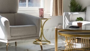 Een moderne woonkamer met een grijze fauteuil, een ronde salontafel met gouden poten, een klein bijzettafeltje met een decoratief object, en een uitzicht op een gebouw door een raam met beige gordijnen.