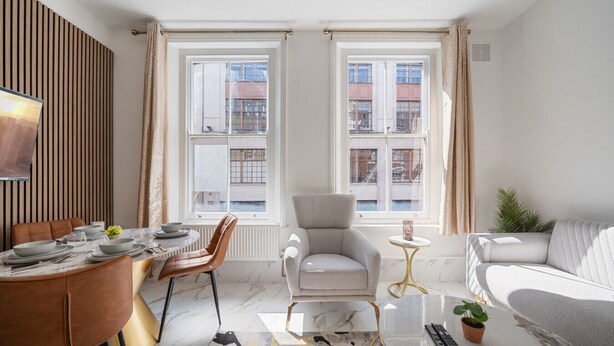 Een moderne woonkamer met een eethoek, grote ramen, een grijze fauteuil, een bank en een kleine tafel met een plant.