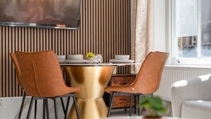 Een eetgedeelte met een ronde gouden tafel, twee bruine stoelen en een stadsgezicht door een raam.