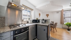 Cocina compacta con estufa a gas, horno y un refrigerador pequeño. Hay una ventana sobre el fregadero, un microondas y una tetera en la encimera. En el fondo se ve una zona de comedor con una mesa y sillas.