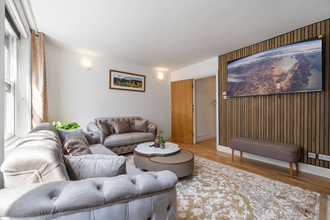 A modern living room with a grey sofa set, a round coffee table, and a large flat-screen TV mounted on a wooden wall.