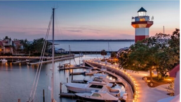 Ein Yachthafen im Abendlicht mit vertäuten Booten, einem Leuchtturm und einem von Bäumen gesäumten Weg.