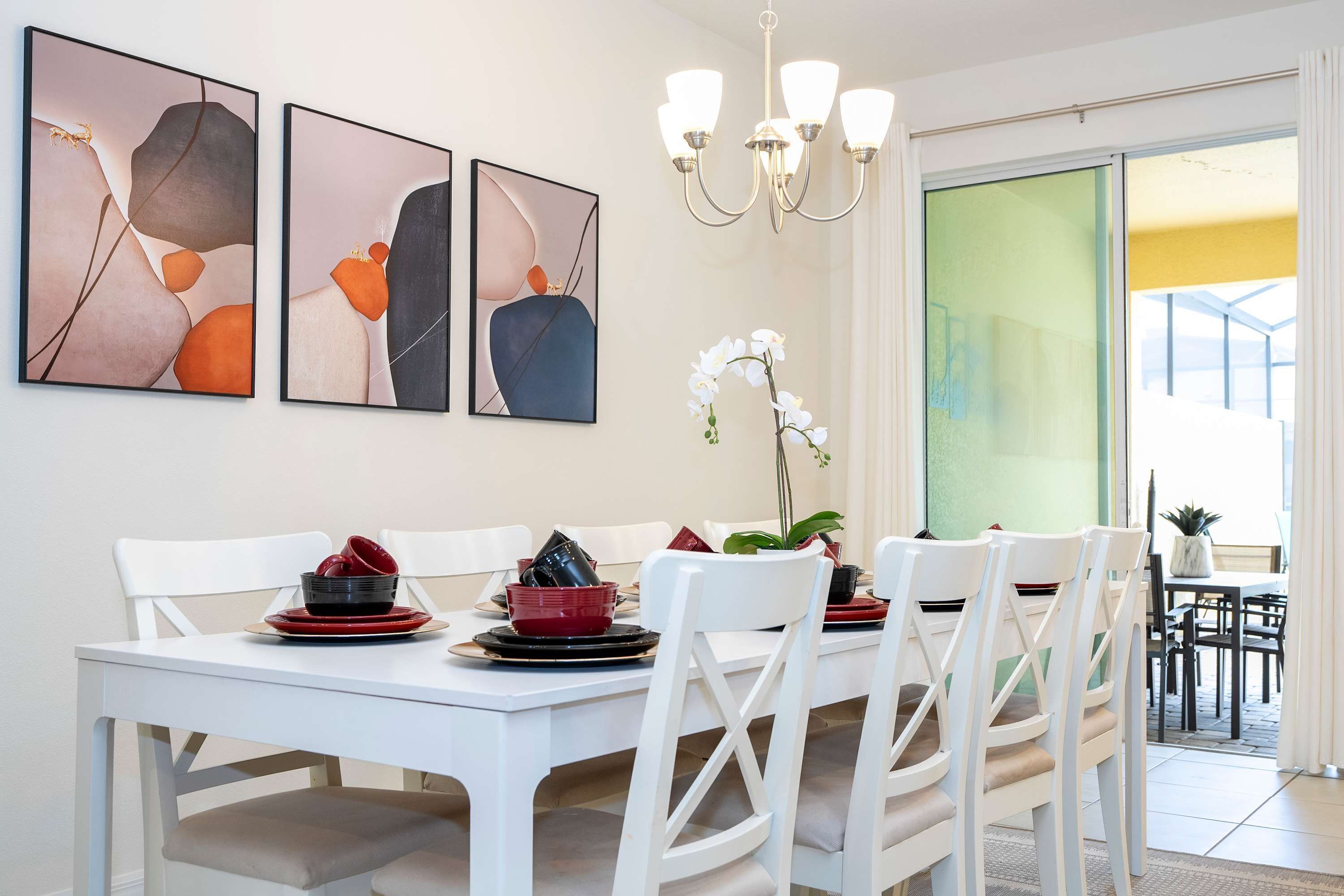 Een eetruimte met een witte tafel gedekt voor vier personen, omringd door witte stoelen. Boven de tafel hangen drie kunstwerken in lijsten.